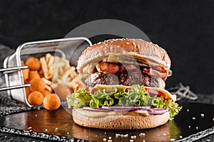 Beef burger with lettuce, tomatoes, slice of cheese, ham, pastrami, sauce and fried stick balls, french fries on slate black