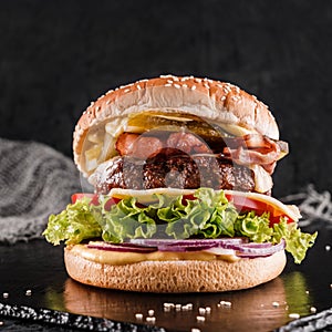 Beef burger with lettuce, tomatoes, slice of cheese, ham, pastrami and sauce on slate black background, close up
