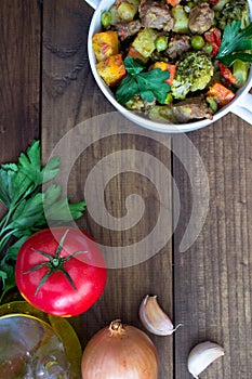 Beef meat stewed with vegetables in ceramic pot with ingredients on wooden background