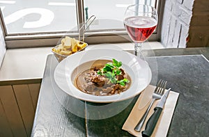 Beef stew or goulash in a delicious rich brown gravy on a white plate with french fries and glass of drink over a window