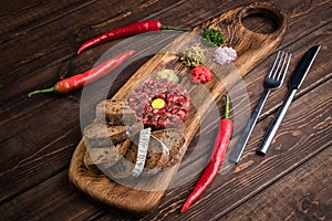 Beef Tartar with a raw egg yolk, rye bread and chilis on a wooden board with spoon and fork. Shot made from above on a dark rustic