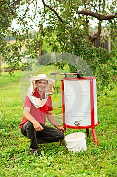 A beekeeper in a protective cap crouched near the honey extractor. He pours fresh honey into the white bucket