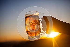 Beer glass in the hand against blue sky and sun in the evening. Outdoor party, picnic concept