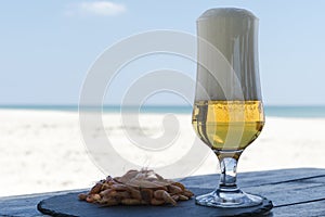 Beer glass and shrimps on stone plate in shadow table against sunny beach blurred background of the sea.