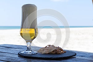 Beer glass and shrimps on stone plate in shadow table against sunny beach blurred background of the sea.