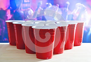 Beer pong tournament layout. Many red party cups in a nightclub full of people dancing on the dance floor in the background.