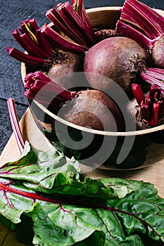 Beets with green tops in round metal pan on dark black wooden background, fresh red beetroot on backdrop kitchen table top view