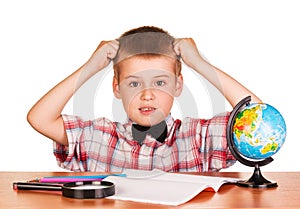 Befuddled boy sitting at table on notebook, pencils, globe .