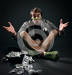 Beggar, pauper man in jacket, jeans and shoes sits cross legged on floor by upside down hat full of dollars cash