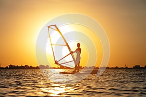 A beginner windsurfer woman stands on a board with a sail on a sunset background. Windsurfing school