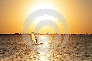 A beginner windsurfer woman stands on a board with a sail on a sunset background. Windsurfing school