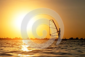 A beginner windsurfer woman stands on a board with a sail on a sunset background. Windsurfing school