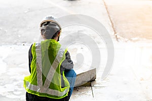 Behind a woman, a construction engineer wearing a hat and a green safety vest, stands looking at work in the construction zone