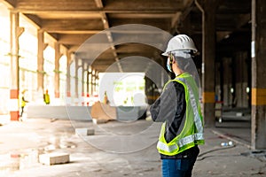 Behind a woman, a construction engineer wearing a hat and a green safety vest, stands looking at work in the construction zone