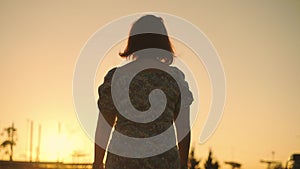 Behind a woman strolling through a wide field behind a wind turbine generating electricity in the evening.