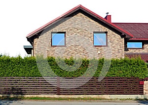 Behind a wooden fence and green coniferous hedge is the side wall of a yellow rural brick house isolated