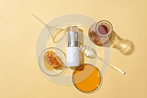 On a beige background, a cosmetic bottle, beeswax on a petri dish, honey in a glass jar, and a honey drizzle form a composition.