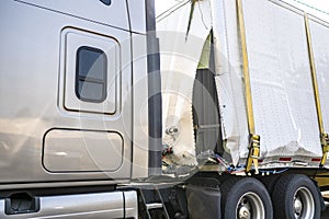 Beige big rig semi truck with a damaged in an accident dry van semi trailer secured with slings for transportation to the repair