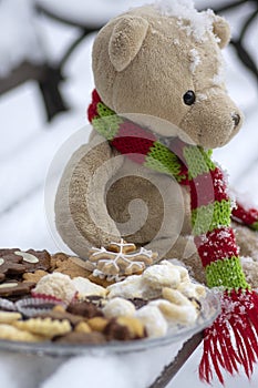 Beige plushy teddy bear with red green striped knitted scarf sitting with Christmas cookies on the bench covered with white snow