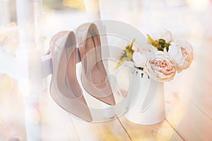 beige shoes for bride hanging on a white chair against a background of a bouquet of pions