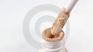 Beige skin concealer pouring from a brush into the bottle on light background close up