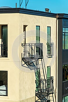 Beige stucco building detail with black metal fire escape ladder and platform with blue sky background