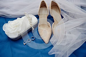 Beige suede female shoes, clutch bag and golden wedding rings on blue background, copy space.