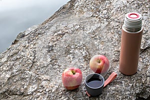 Beige thermos bottle, cup of tea or coffee, and red apple on rock over the river. Beautiful summer, spring, autumn landscape.