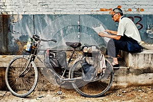 Man reading a newspaper next to his bicycle in a typical city hutong