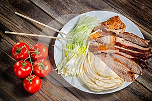 Beijing chinese Duck meat served with onions, pancakes, cucumber pieces and tomatoes on white plate with chopsticks close up. Wood