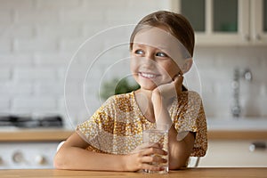 Healthy small girl dreaming by table holding glass of water