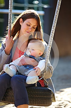 Being a mom can be so gratifying. Young mom on a park swing with her baby boy.