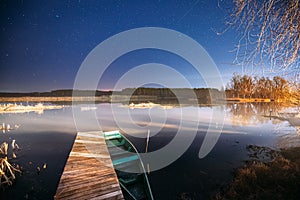 Belarus, Eastern Europe. Real Night Sky Stars Above Old Pier With Moored Wooden Fishing Boat. Natural Starry Sky And