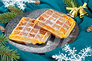 Belgian waffles on a black plate sprinkled with icing sugar. Snowflakes, cones, pine branches on turquoise fabric