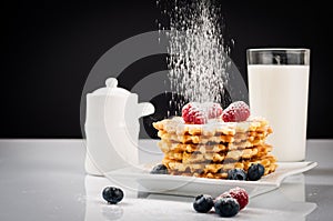 Belgian waffles with raspberries and sieving sugar powder and honey served with jug of milk on a white table