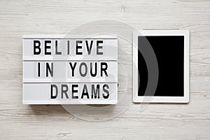 `Believe in your dreams` word on modern board, tablet with blank screen over white wooden background, overhead view. Top view, f