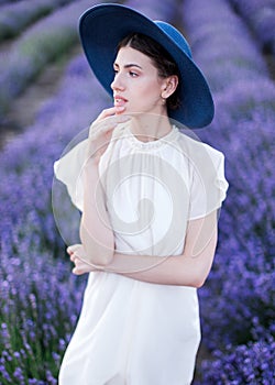 A belted portrait of a beautiful girl in a white dress and a blue hat standing in the middle of blossoming lavender fields and