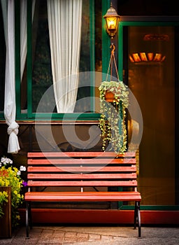 A bench against the wall of the house, in the evening, illuminated by the light of a street lamp. Flowerpot, flowerbed, flowers.