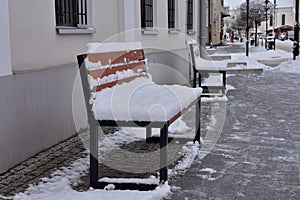 A bench covered with snow against the wall of a building in a city on a gloomy winter day. Winter