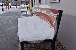 A bench covered with snow against the wall of a building in a city on a gloomy winter day. Winter