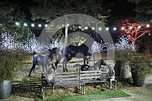 Bench decorated with statues and three bronze horses in the back of the bench at a park lit with colorful light orna