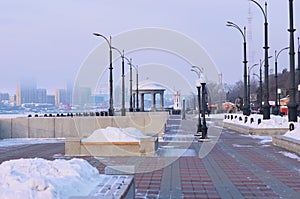 A bench with a flower bed in winter, filled with a snowdrift. Rotunda and lighthouse on the embankment. Blurred in the