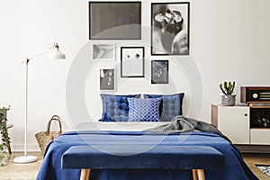 Bench in front of bed with navy blue pillows between lamp and cabinet in bedroom interior. Real photo