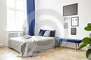 Bench next to bed with grey blanket in bright bedroom interior with posters and blue drapes. Real photo