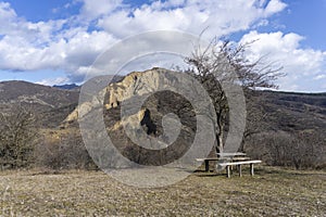A bench with a table in a clearing under a tree overlooking a partially destroyed mountain. Rock formations are visible. There are