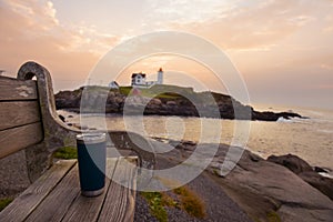 A bench and a thermos of coffee with a view of the sunrise on the coast and a lighthouse . USA. Maine. Nubble Lighthouse