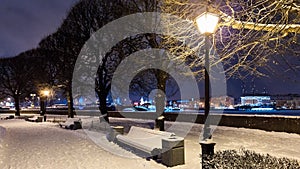 Benches under snow along alley with trees and street lights on embankment. Winter cityscape at twilight. Saint Petersburg, Russia.