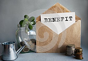 BENEFIT text is written on white paper on an antique envelope, which lies on the table along with a stack of coins, a glass jar of