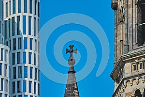 Pointed tower with cross of the memorial church in front of a blue sky next to a modern high-rise building with strict rectangular