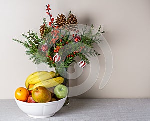 Berries pine cones and fresh winter greenery as a bouquet on a white table with fruit in thw vase.